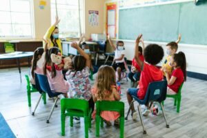 Diverse group of children in a classroom, raising hands, led by masked teacher. Bright and engaging learning environment.